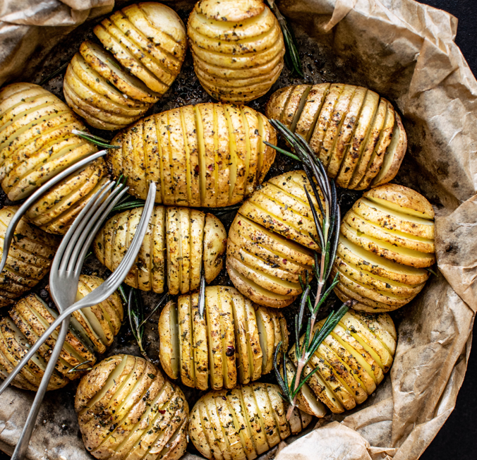 Gebackene Kartoffeln mit eingeschnittenen Oberflächen in einer Schale auf Backpapier, dekoriert mit Rosmarinzweigen, daneben eine Gabel und ein Messer.