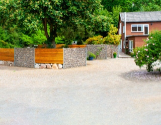 Backsteingebäude mit grauem Dach, umgeben von Bäumen und Sträuchern, mit einer Steinmauer und Holzelementen im Vordergrund.