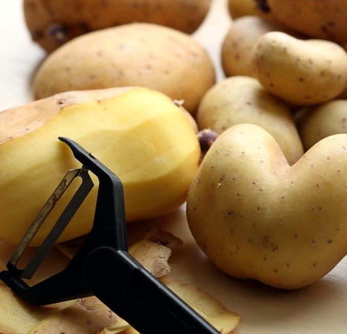 Mehrere Kartoffeln, eine teilweise geschälte Kartoffel, ein Sparschäler und Kartoffelschalen auf einer Oberfläche.