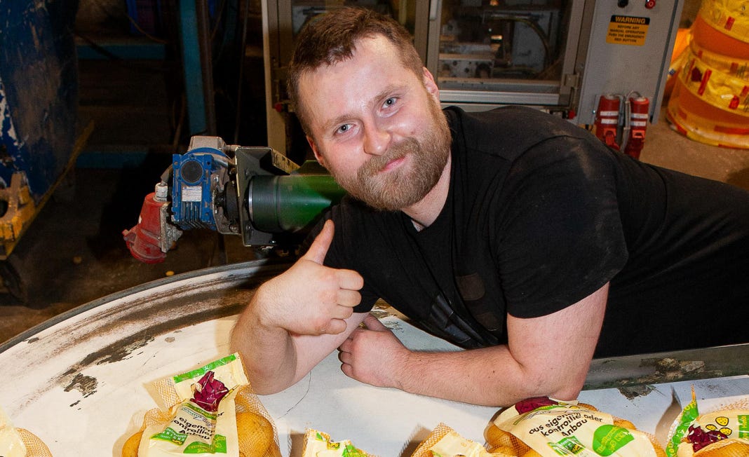 Mann mit Daumen hoch neben mehreren Tüten mit Kartoffeln in einer Fabrikumgebung.