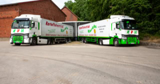Zwei Lastwagen mit grüner und weißer Beschriftung stehen nebeneinander zur Beladung auf einem gepflasterten Platz vor einem roten Backsteingebäude und Bäumen im Hintergrund.