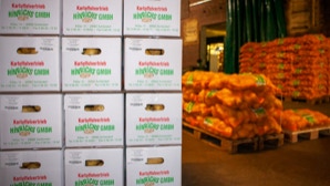 Gestapelte Kisten mit der Aufschrift "Kartoffelvertrieb Hinrichs" neben Paletten mit Säcken voller Kartoffeln in einem Lagerraum.