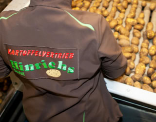 Eine Person in brauner Jacke steht vor einem Förderband voller Kartoffeln.