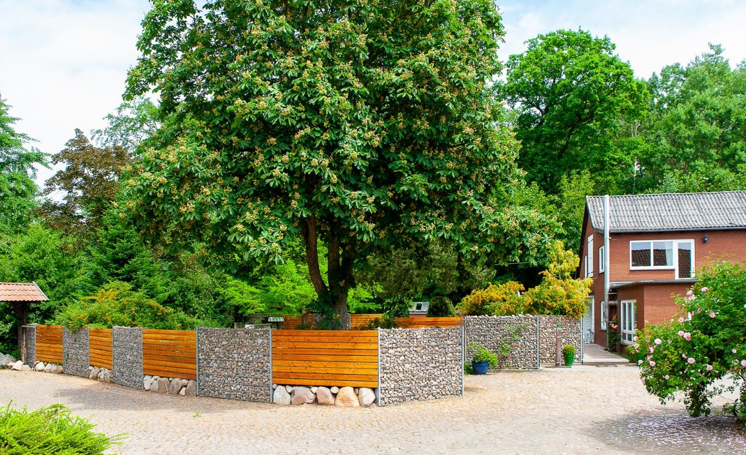 Ein großes Haus mit roter Fassade und grauem Dach, umgeben von Bäumen und einem Garten mit Stein- und Holzzäunen.