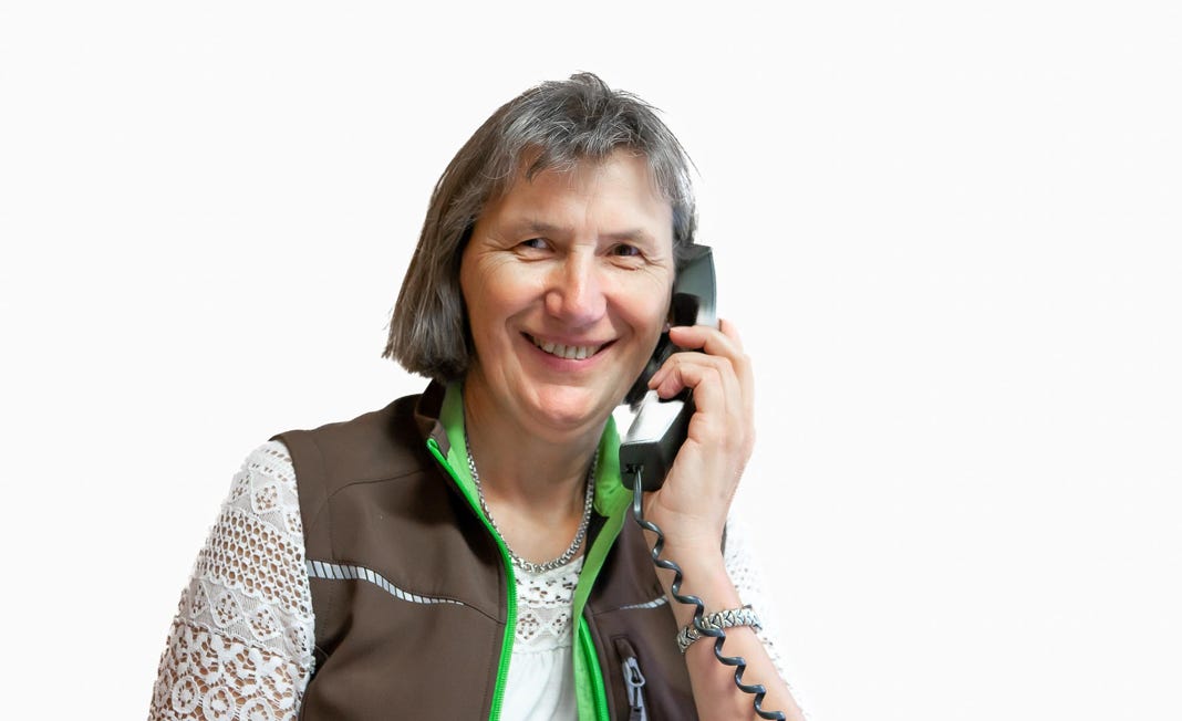 Eine lächelnde Person hält einen Telefonhörer ans Ohr, trägt eine braune Weste und ein weißes Spitzenoberteil.