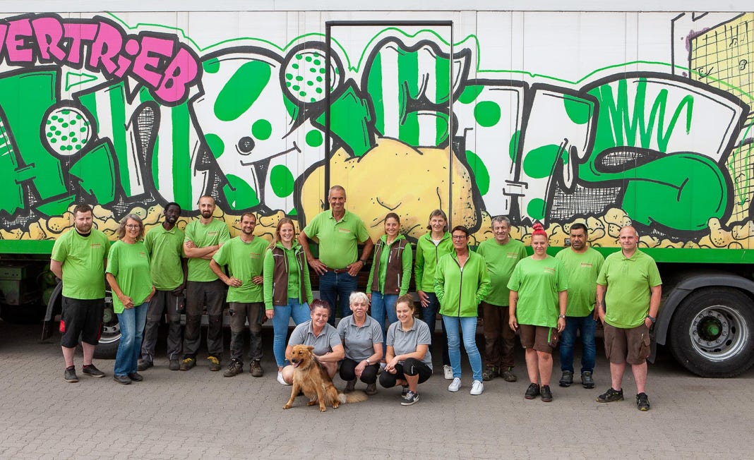 Eine Gruppe von Menschen in grünen T-Shirts steht vor einem bunt bemalten Lastwagen mit Graffiti. Ein Hund sitzt vorne bei einigen Personen, die auf dem Boden hocken.